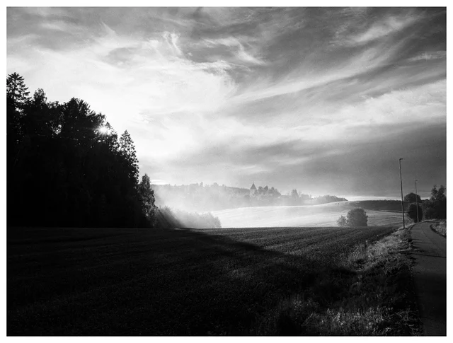 Black and white landscape with sun rays breaking through trees onto misty farmland, a distant church, and a country road beneath dramatic clouds.