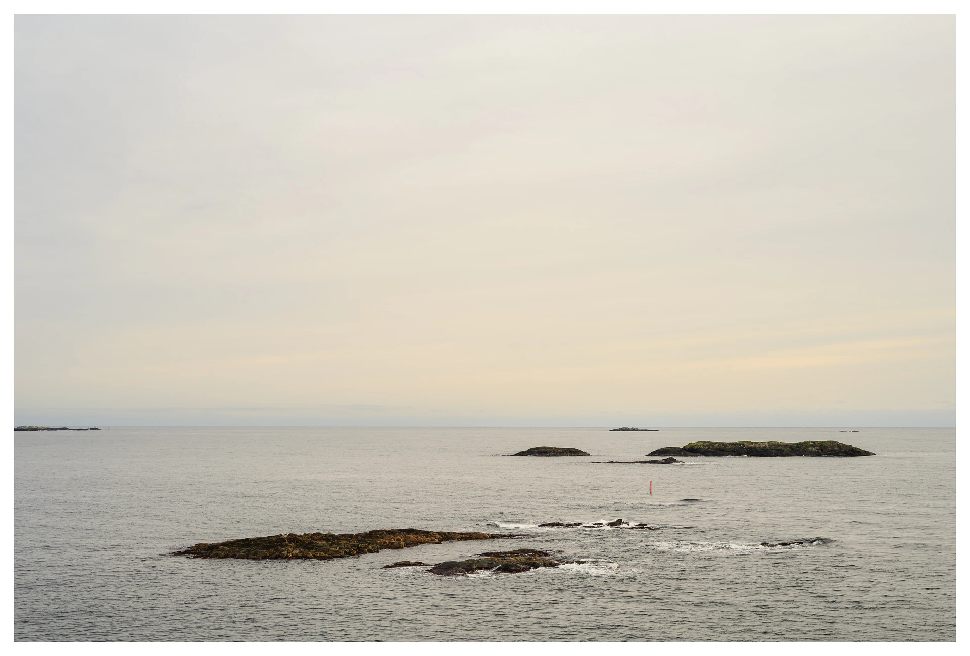Minimal coastal scene with grey water, low rocky islands, and a pale overcast sky stretching to the horizon.
