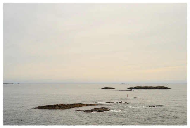 Minimal coastal scene with grey water, low rocky islands, and a pale overcast sky stretching to the horizon.
