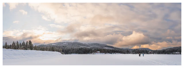 Winter panorama of a frozen lake with cross-country skiers, snow-covered forest, and soft pastel clouds over rolling hills.