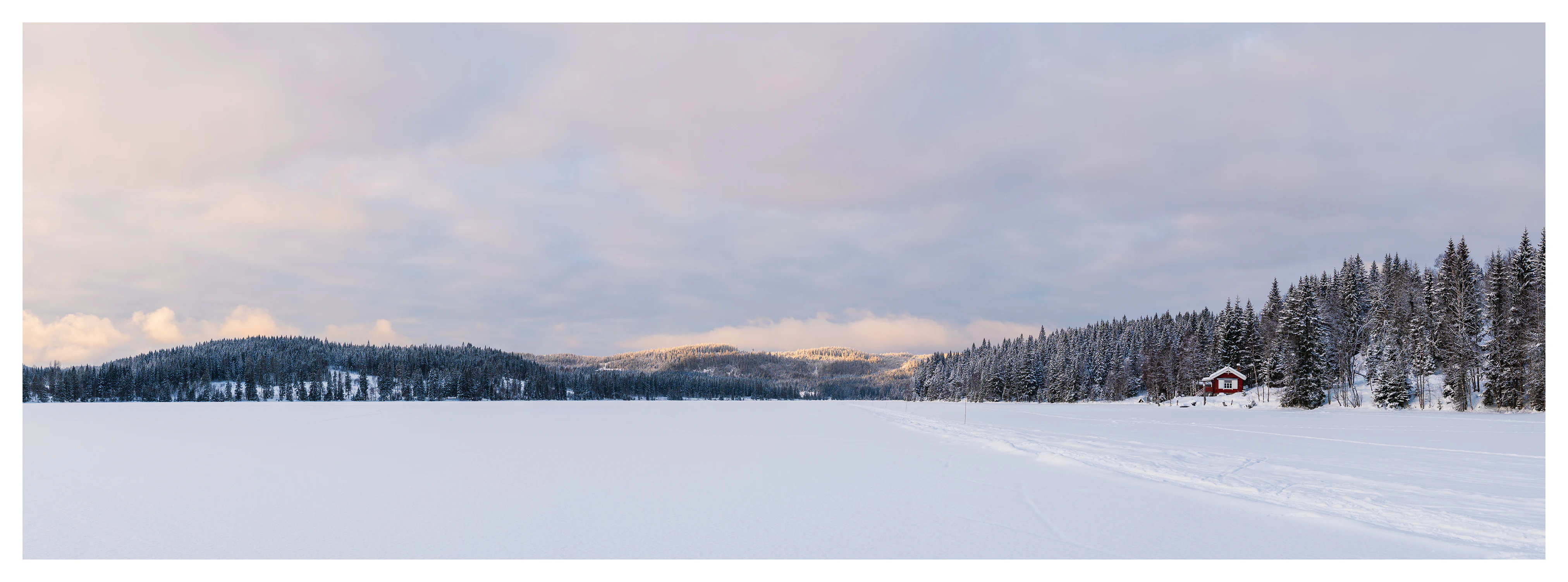Panoramic winter landscape of a frozen lake with snow-covered forest, distant hills, ski tracks, and a red cabin along the shoreline.