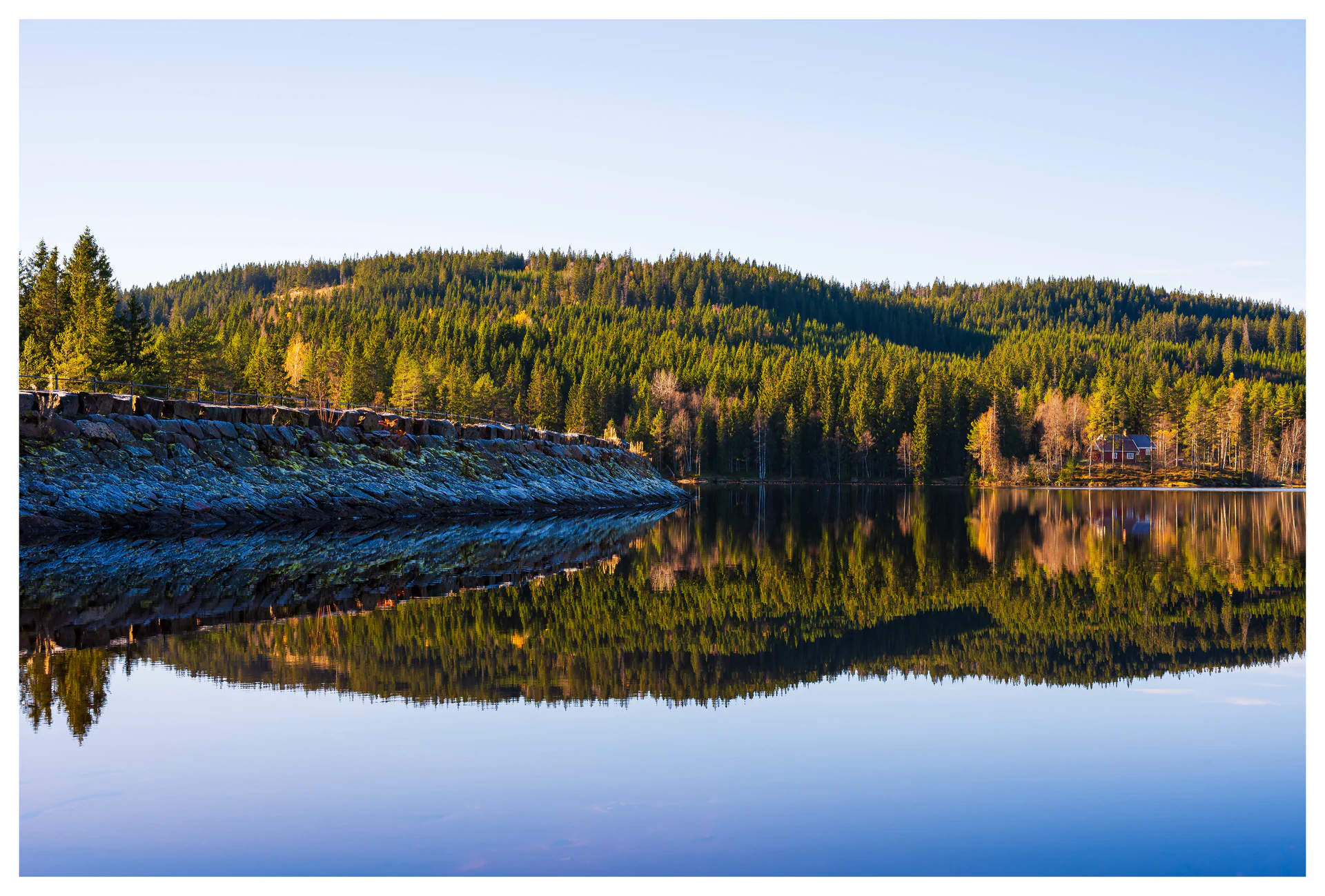 Forest-covered hills and a stone dam reflected in the calm waters of Ørfiske lake in Nordmarka, Norway