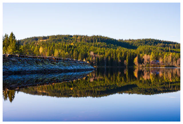 Forest-covered hills and a stone dam reflected in the calm waters of Ørfiske lake in Nordmarka, Norway