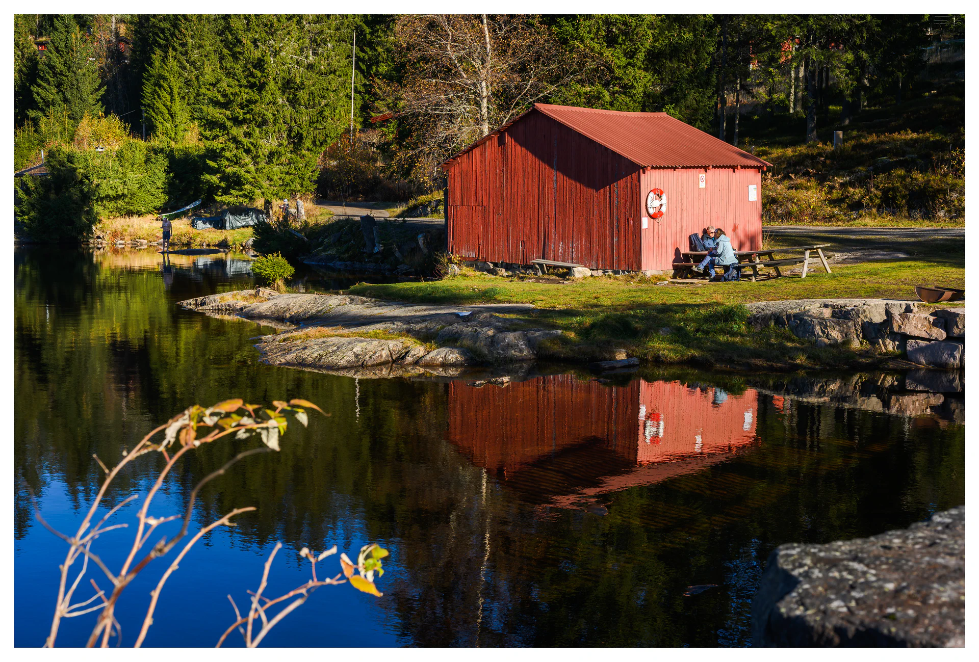 Red boathouse by Ørfiske lake with forest reflections and people resting at a picnic table in Nordmarka, Norway