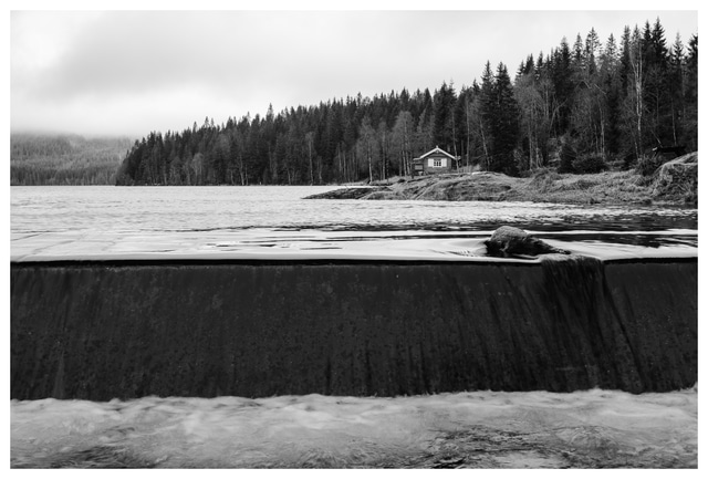 Black and white photograph of the dam at Ørfiske lake, with flowing water in the foreground and a forest cabin in Nordmarka, Norway