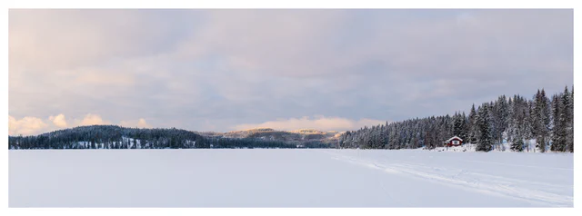 Panoramic winter landscape of a frozen lake with snow-covered forest, distant hills, ski tracks, and a red cabin along the shoreline.