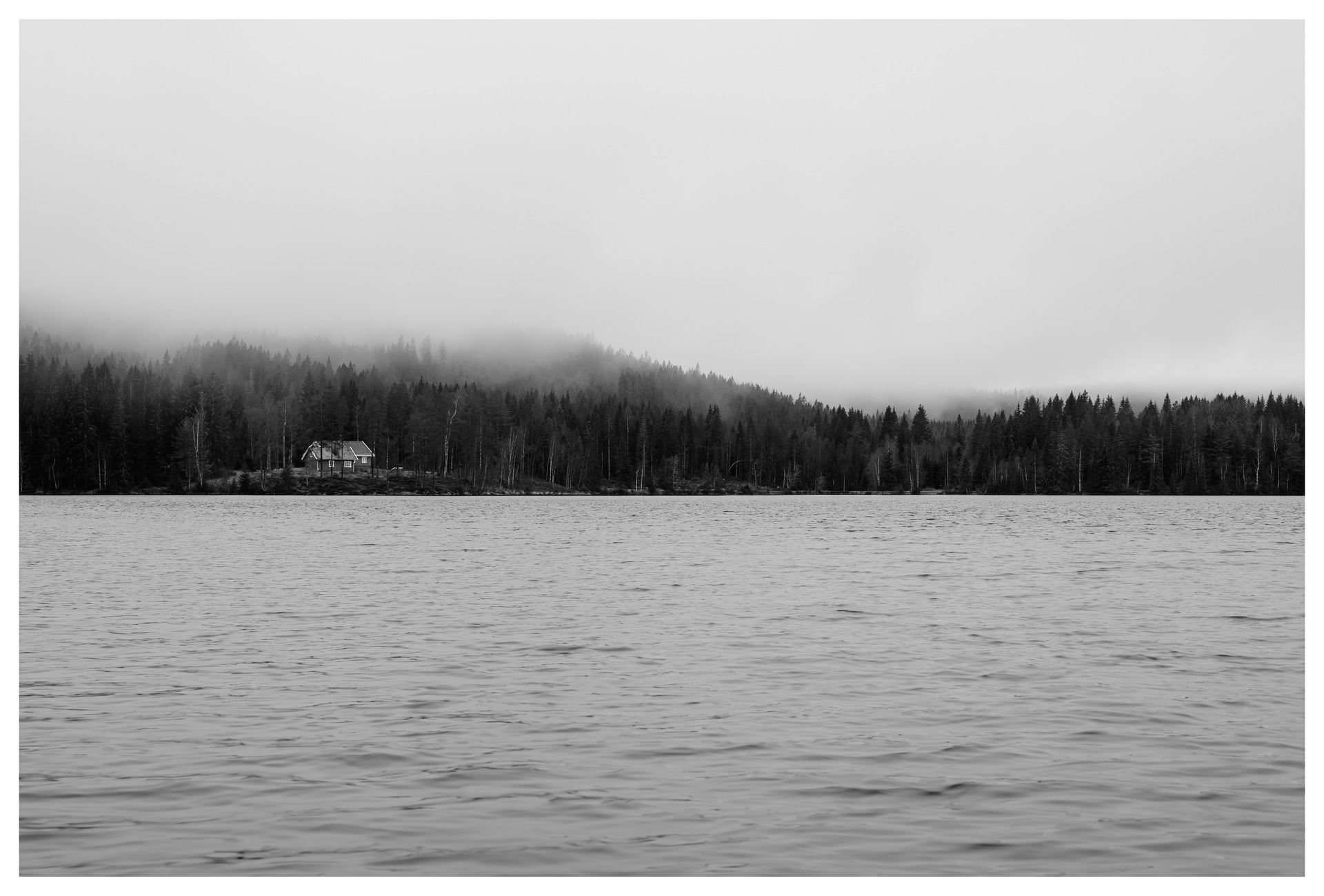 Black and white photograph of Ørfiske lake with a distant forest cabin and misty tree-covered hills in Nordmarka, Norway