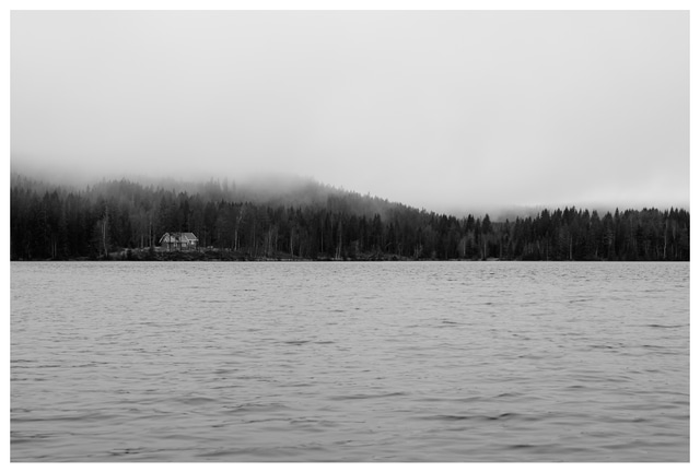 Black and white photograph of Ørfiske lake with a distant forest cabin and misty tree-covered hills in Nordmarka, Norway