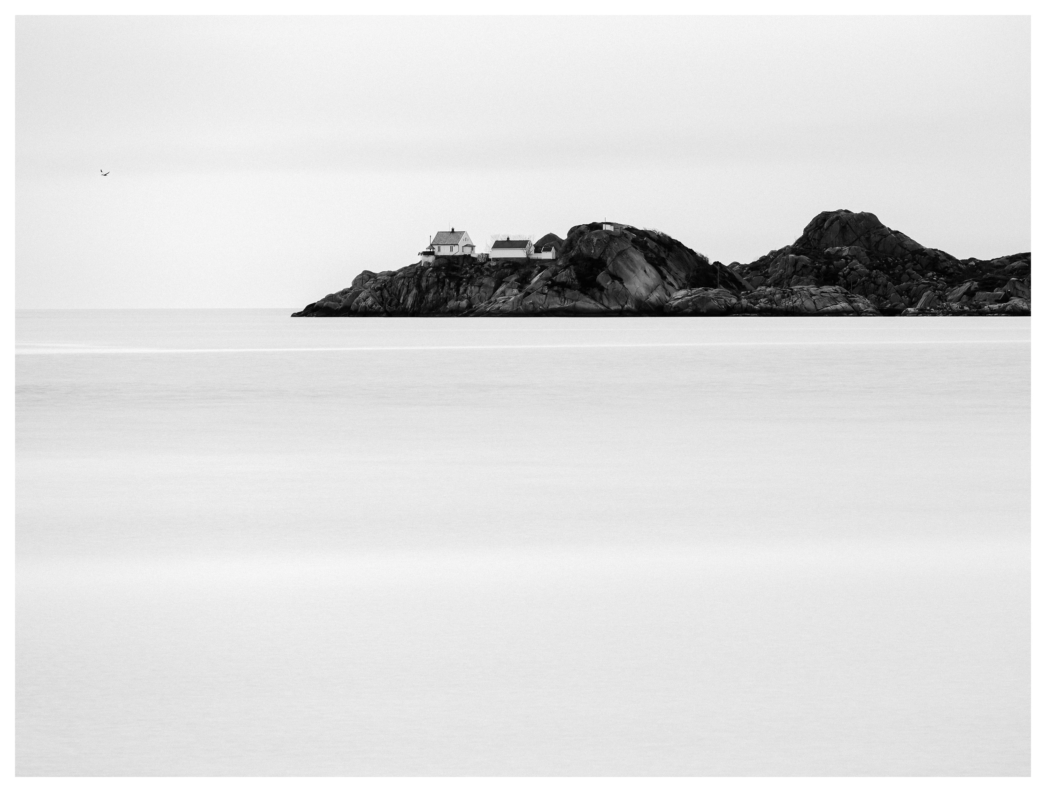 Minimalist black and white photo of a remote house on a rocky island surrounded by calm sea.