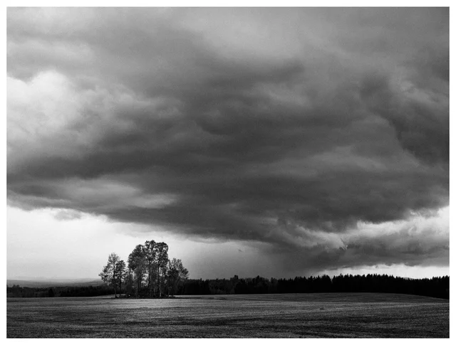Dark storm clouds gathering above an open field and a small tree-covered burial mound near Kløfta, Norway, captured in a dramatic black-and-white landscape.