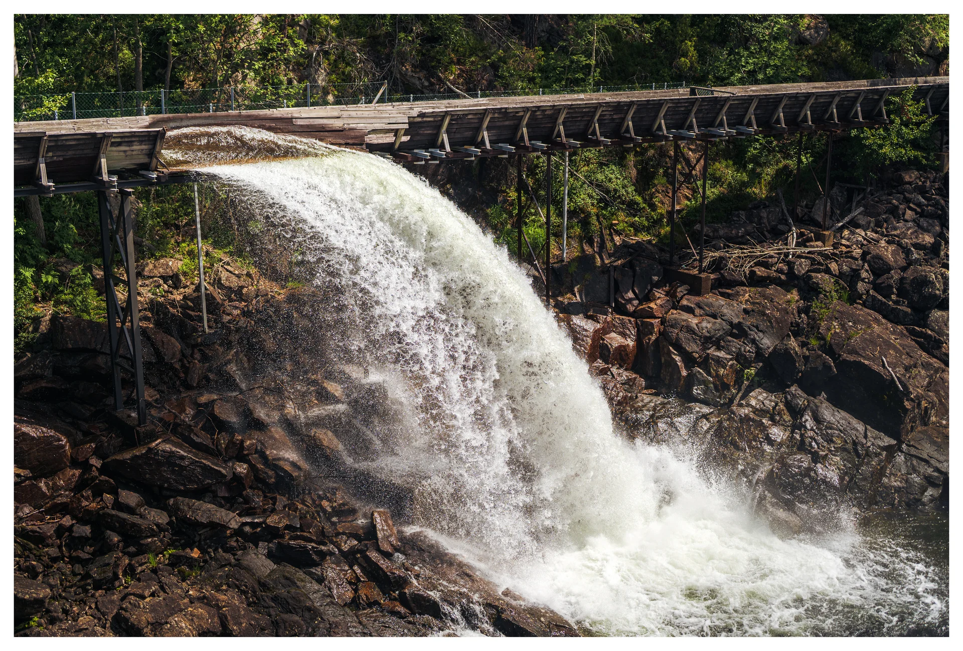 Water pouring over the wooden timber slide in Vennesla, cascading down rocky terrain surrounded by forest.