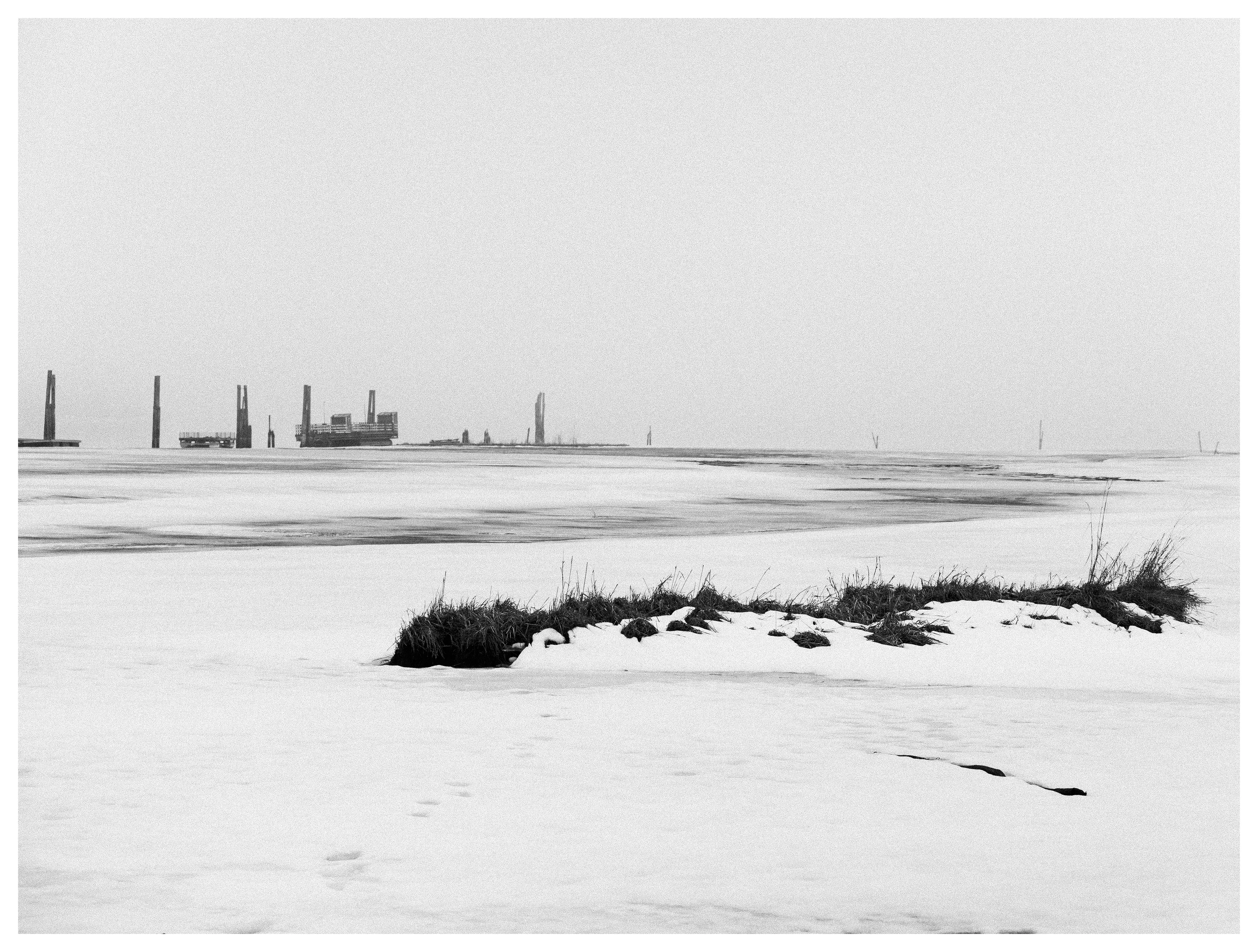 Old timber floating structures from Fetsund Lenser standing in the frozen Glomma River, minimalist winter landscape in black and white with snow and distant wooden pillars.