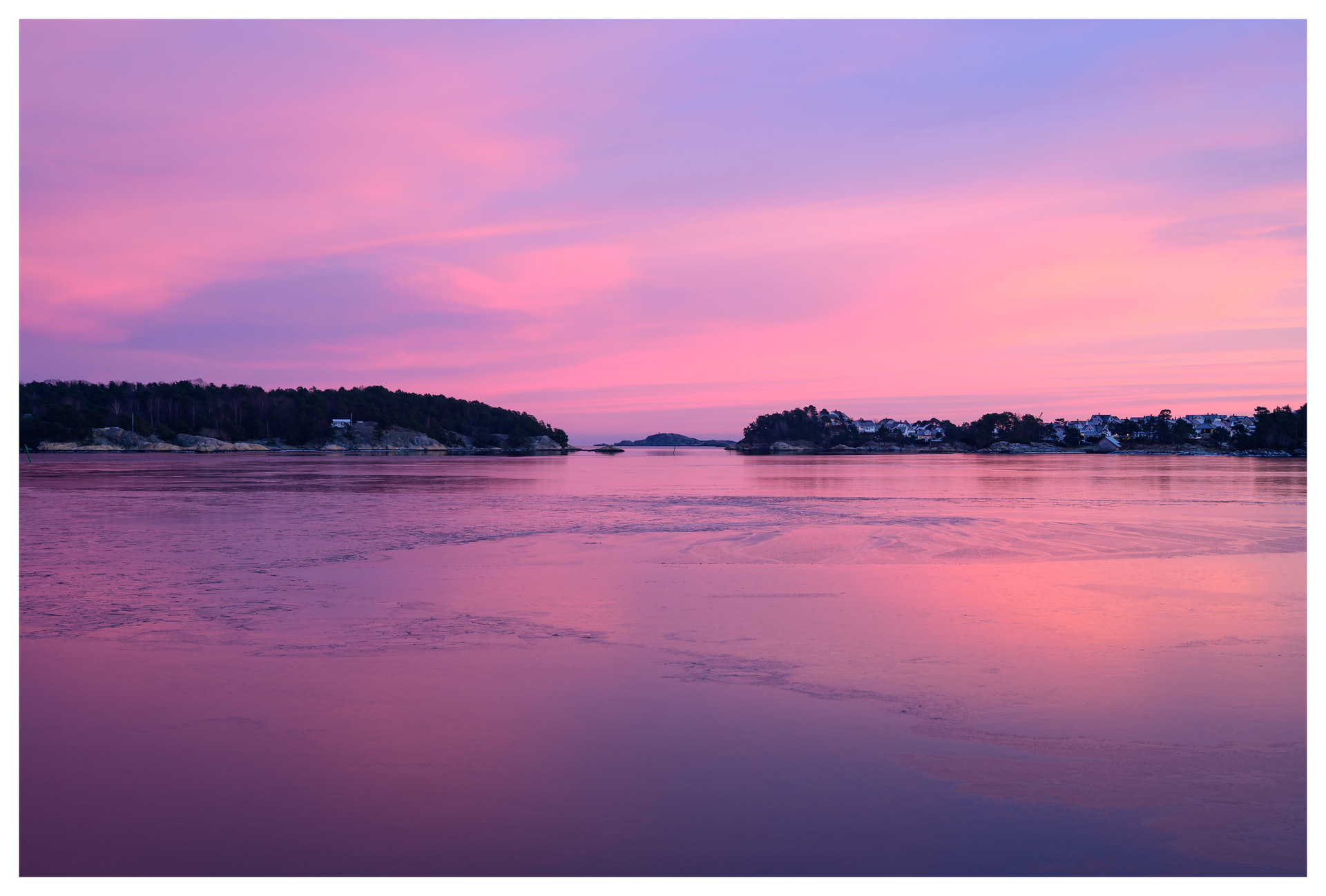 Soft pink and purple winter sky reflected on calm coastal water, with distant islands and shoreline fading into the horizon.