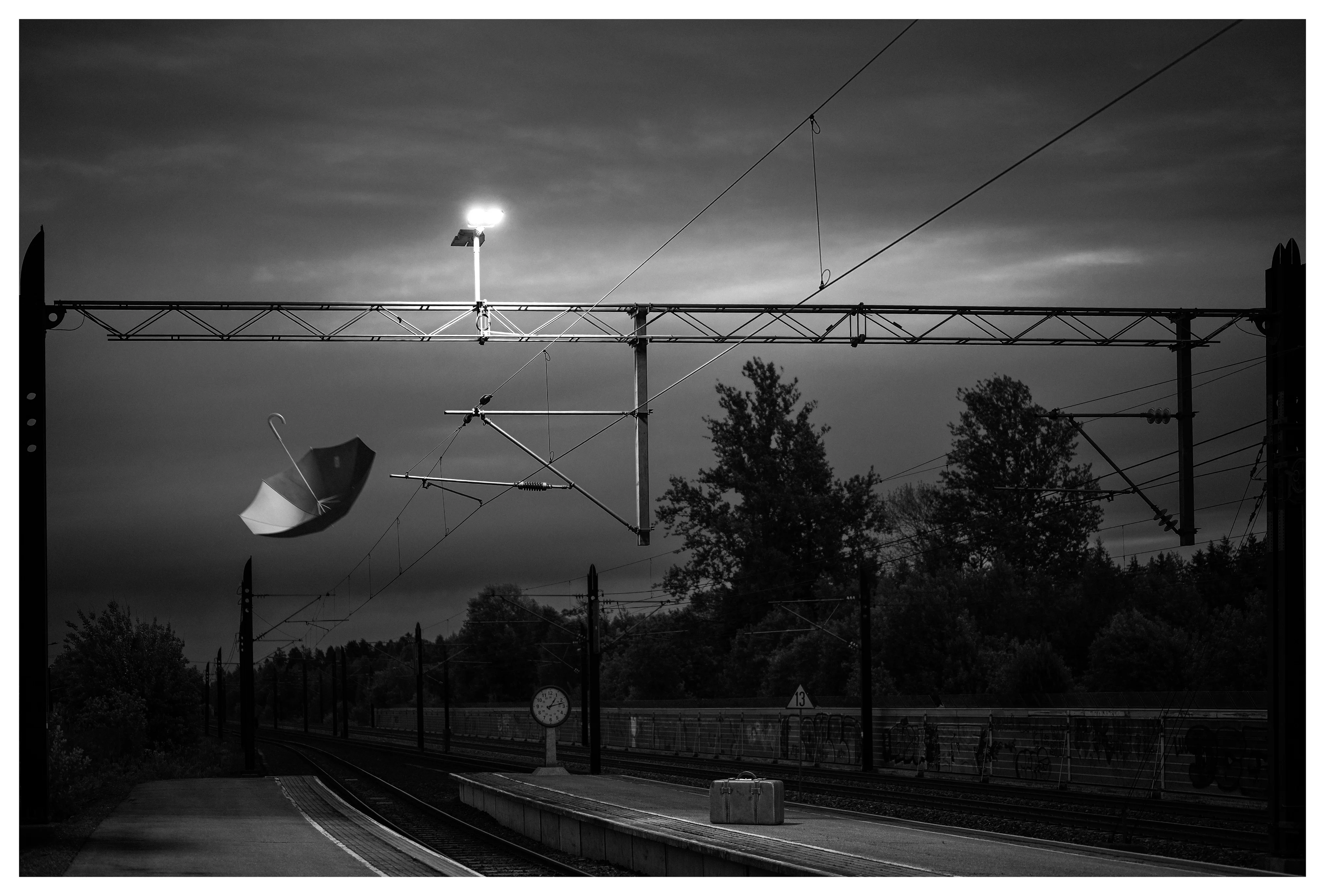 Black-and-white photo of an empty railway platform with overhead power lines and a bright lamp, a suitcase on the platform and a surreal floating umbrella in the sky.