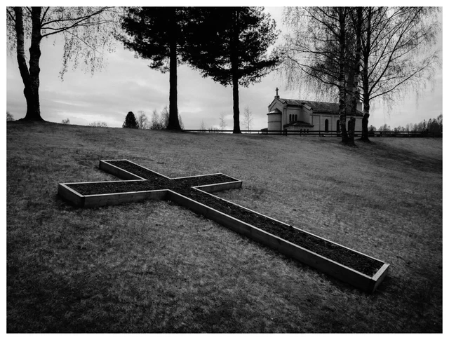 Wooden cross-shaped flower bed on a grassy hillside with a small church in the background, photographed in early spring before bloom.