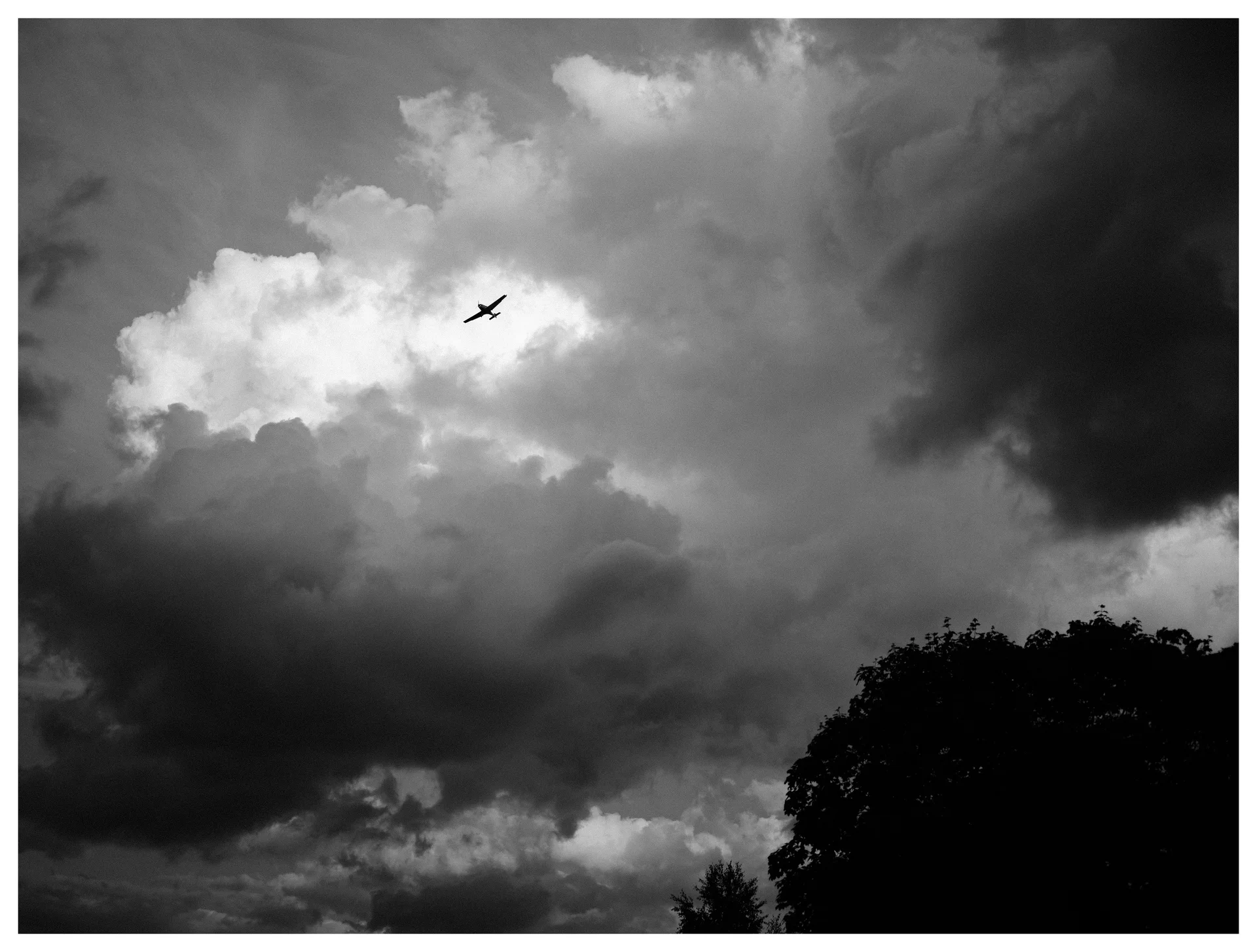 Black and white photograph of an airplane silhouetted against dramatic storm clouds above dark tree silhouettes.