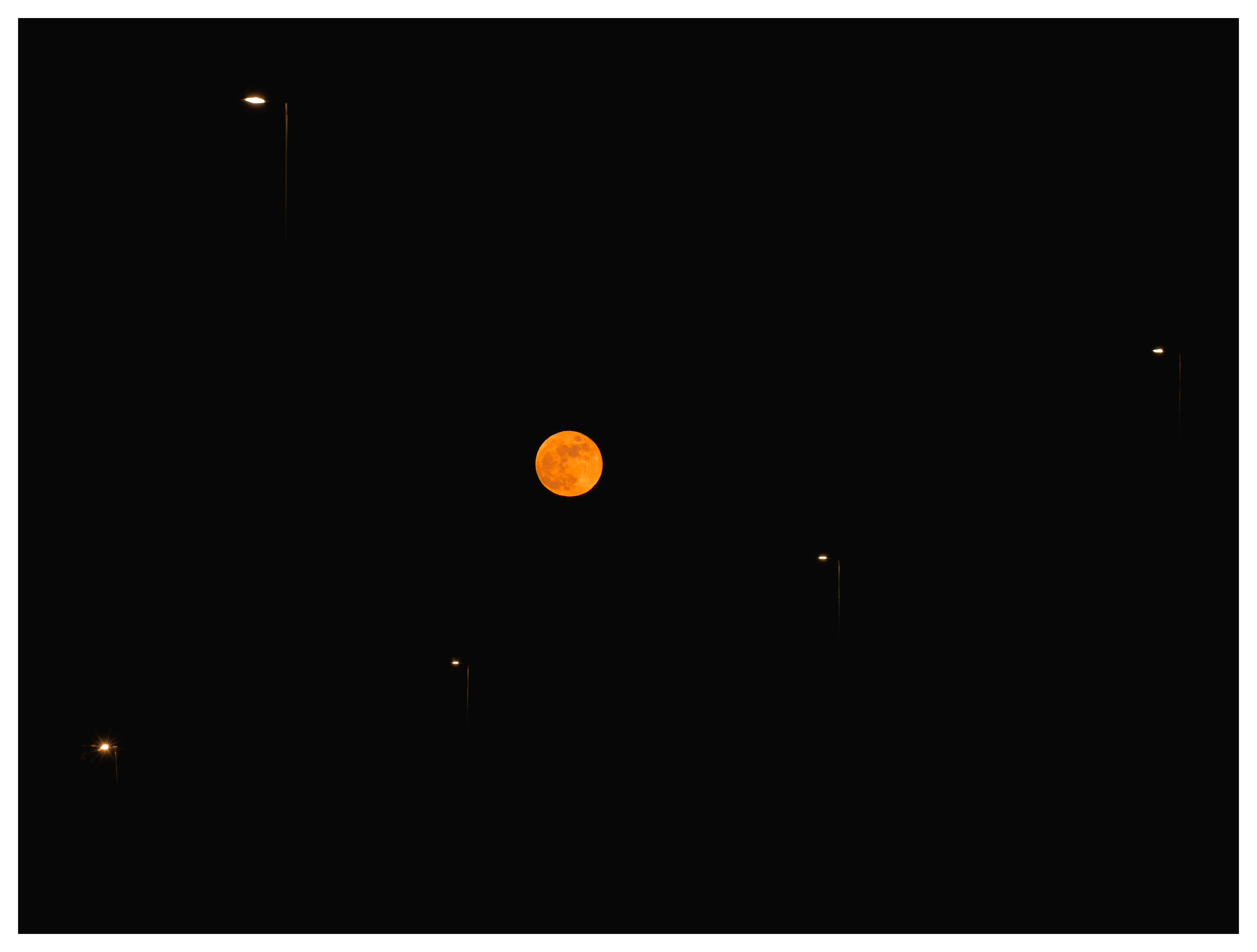 Blood moon during the lunar eclipse on March 3, 2026, glowing deep orange in a black night sky, framed between illuminated streetlights in an urban landscape.