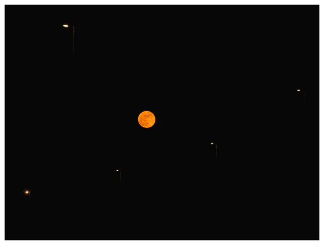 Blood moon during the lunar eclipse on March 3, 2026, glowing deep orange in a black night sky, framed between illuminated streetlights in an urban landscape.