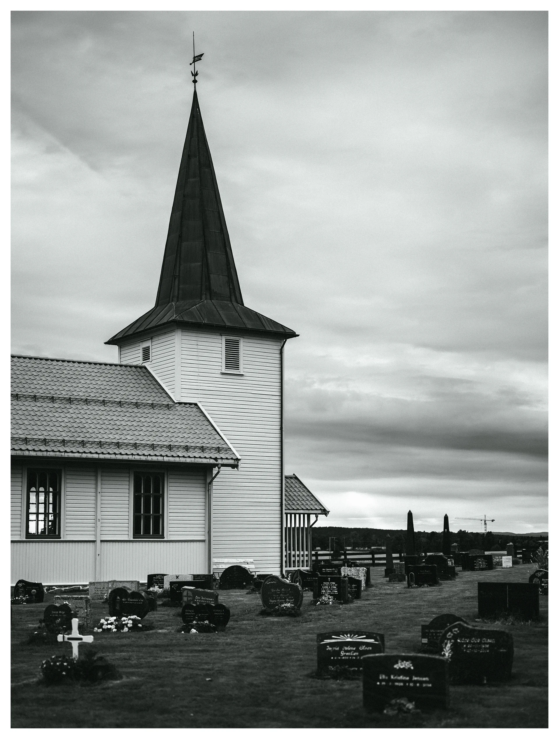 Black and white photograph of a rural church and cemetery beneath a dramatic sky, with a distant construction crane beyond the gravestones.