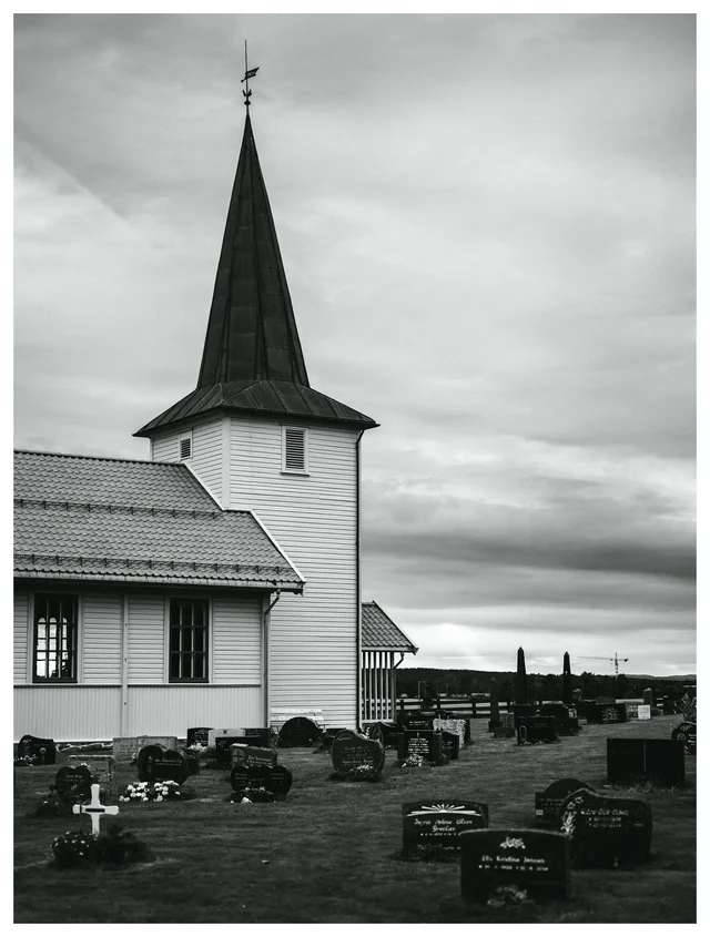 Black and white photograph of a rural church and cemetery beneath a dramatic sky, with a distant construction crane beyond the gravestones.