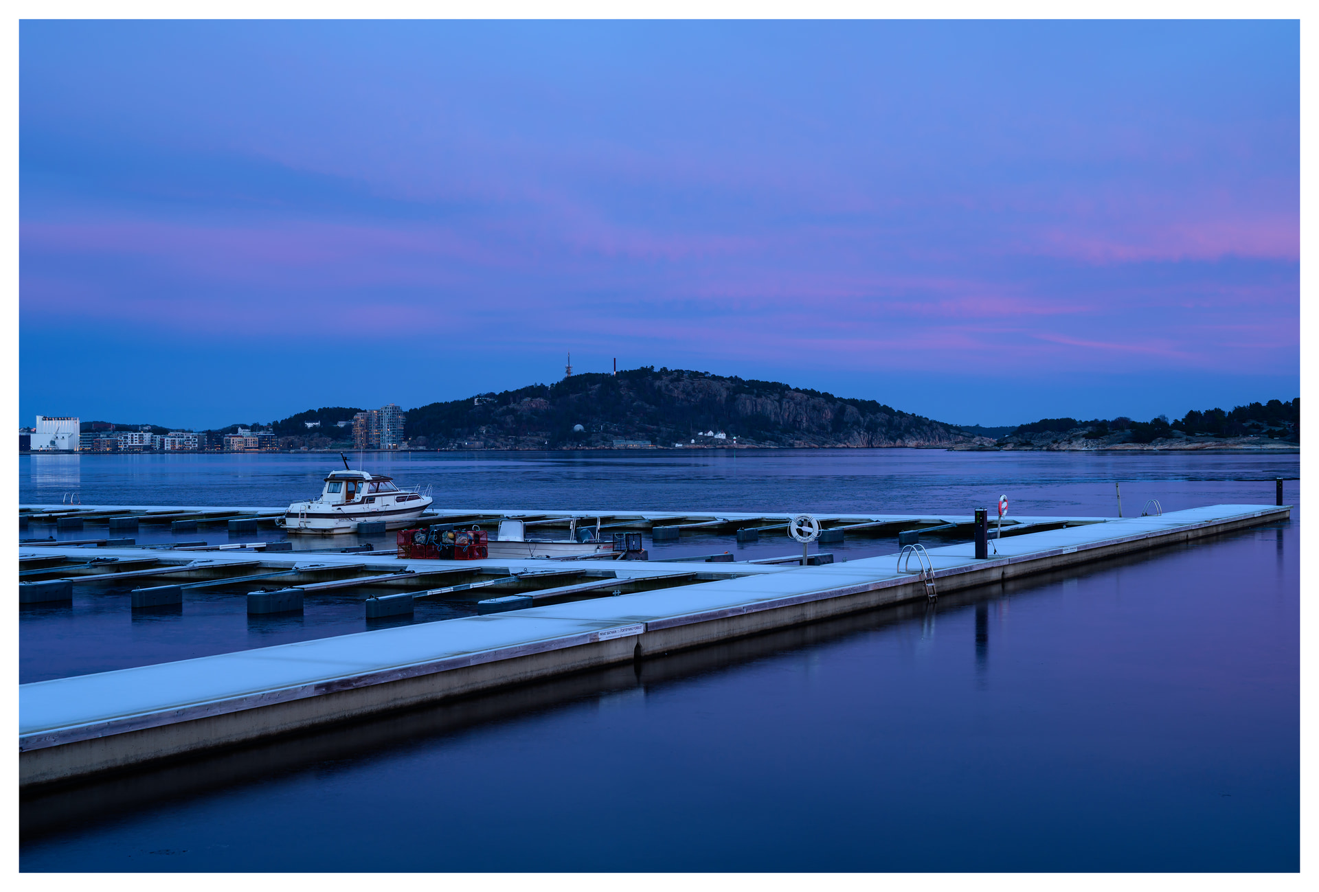 Snow-covered marina docks at blue hour, with a single boat and distant hills under a soft purple winter sky.