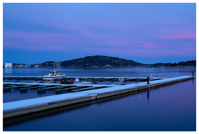 Snow-covered marina docks at blue hour, with a single boat and distant hills under a soft purple winter sky.