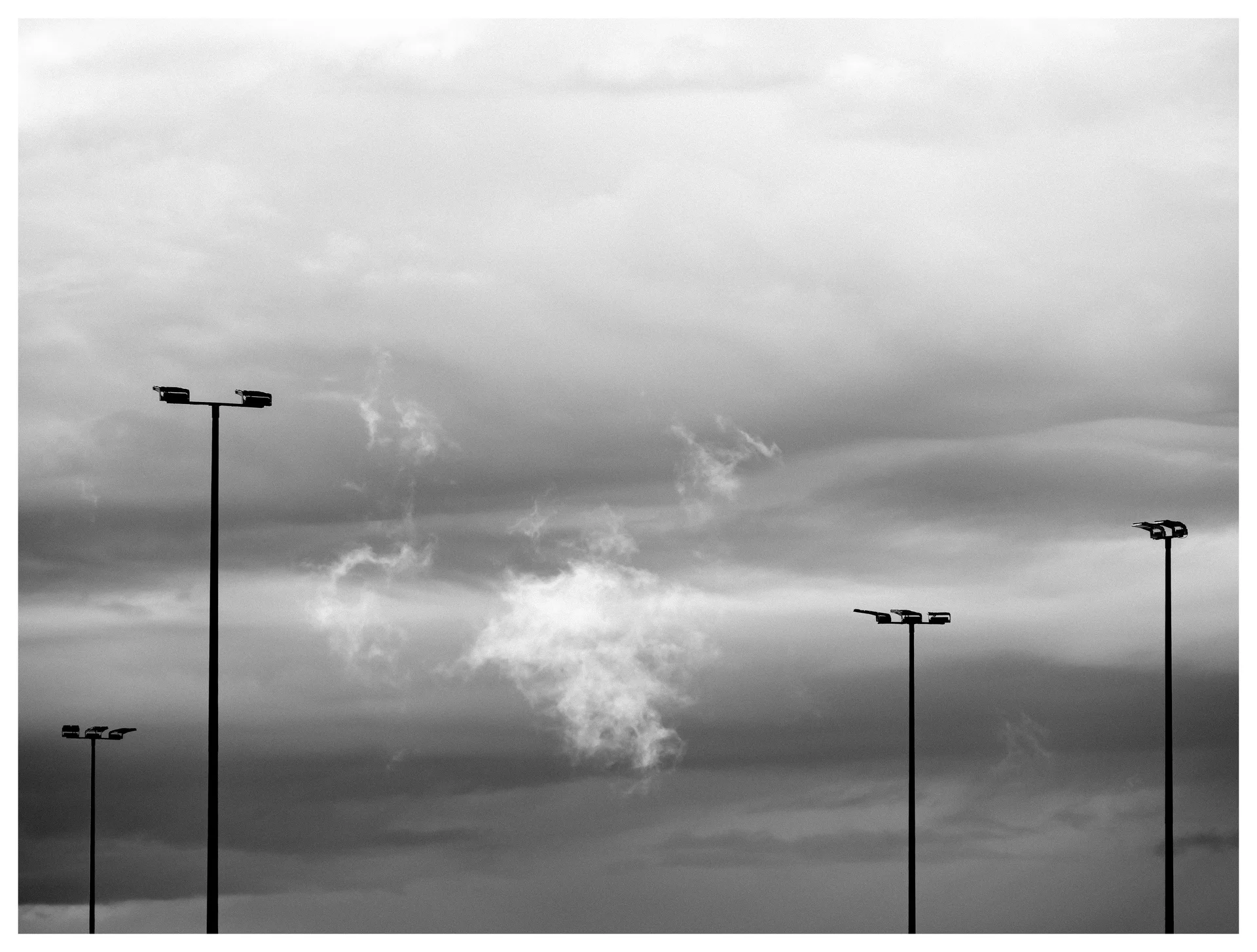 Black-and-white photograph of drifting mist illuminated after rainfall, rising between tall streetlights beneath layered storm clouds.