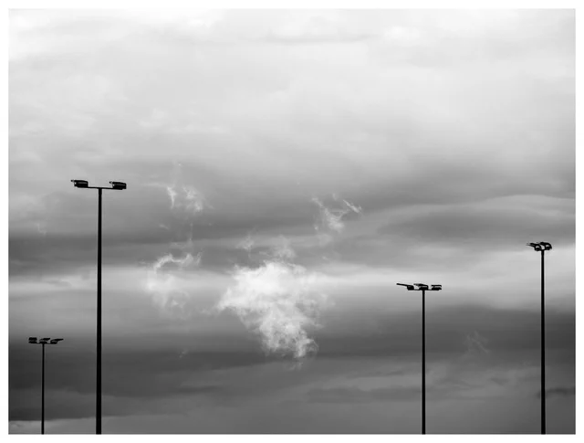 Black-and-white photograph of drifting mist illuminated after rainfall, rising between tall streetlights beneath layered storm clouds.
