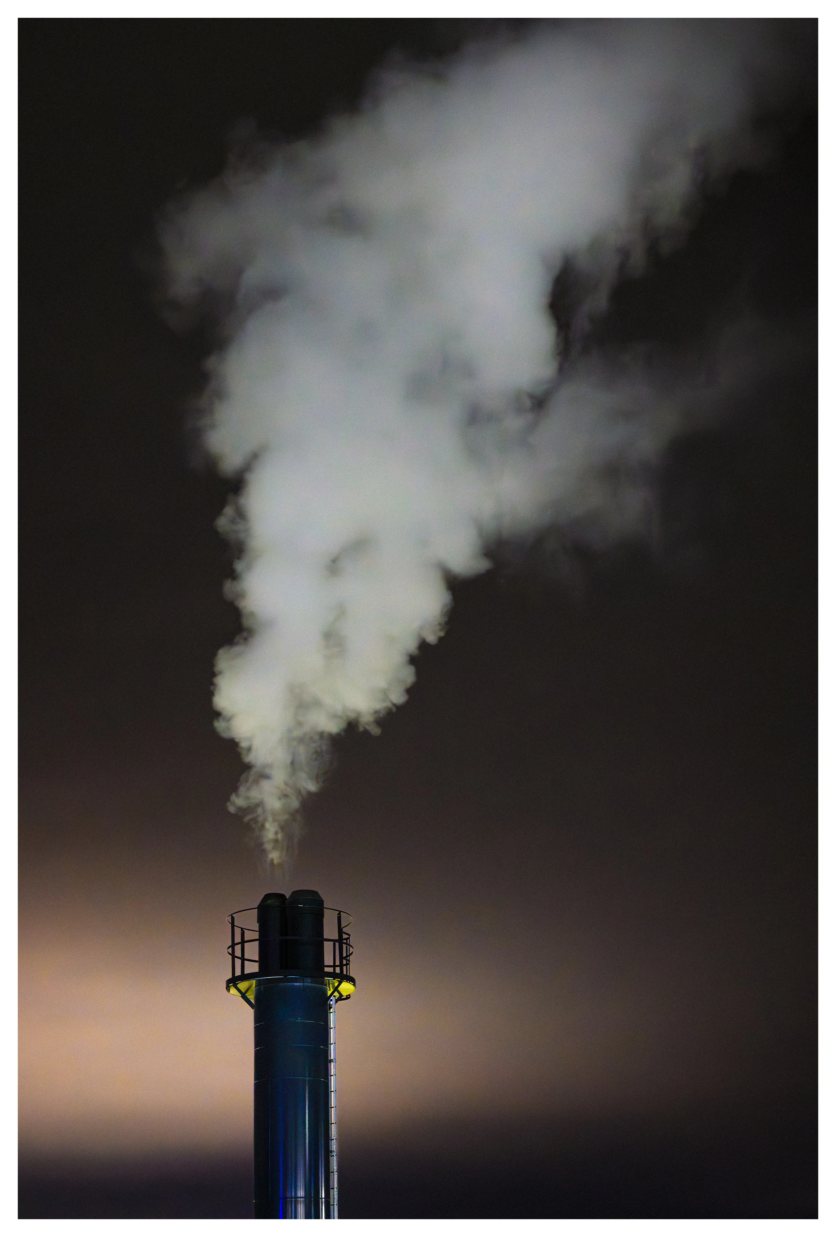 Industrial chimney releasing steam into the night sky, backlit by warm light and surrounded by darkness.