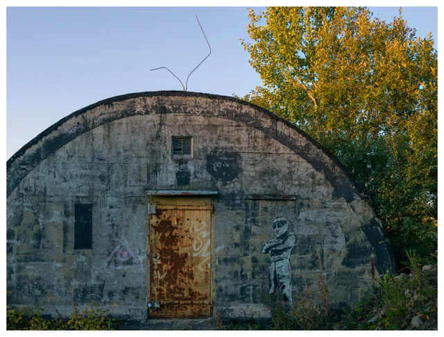 Weathered concrete bunker with a rusted door, graffiti figure and exposed metal rods rising from the curved roof.