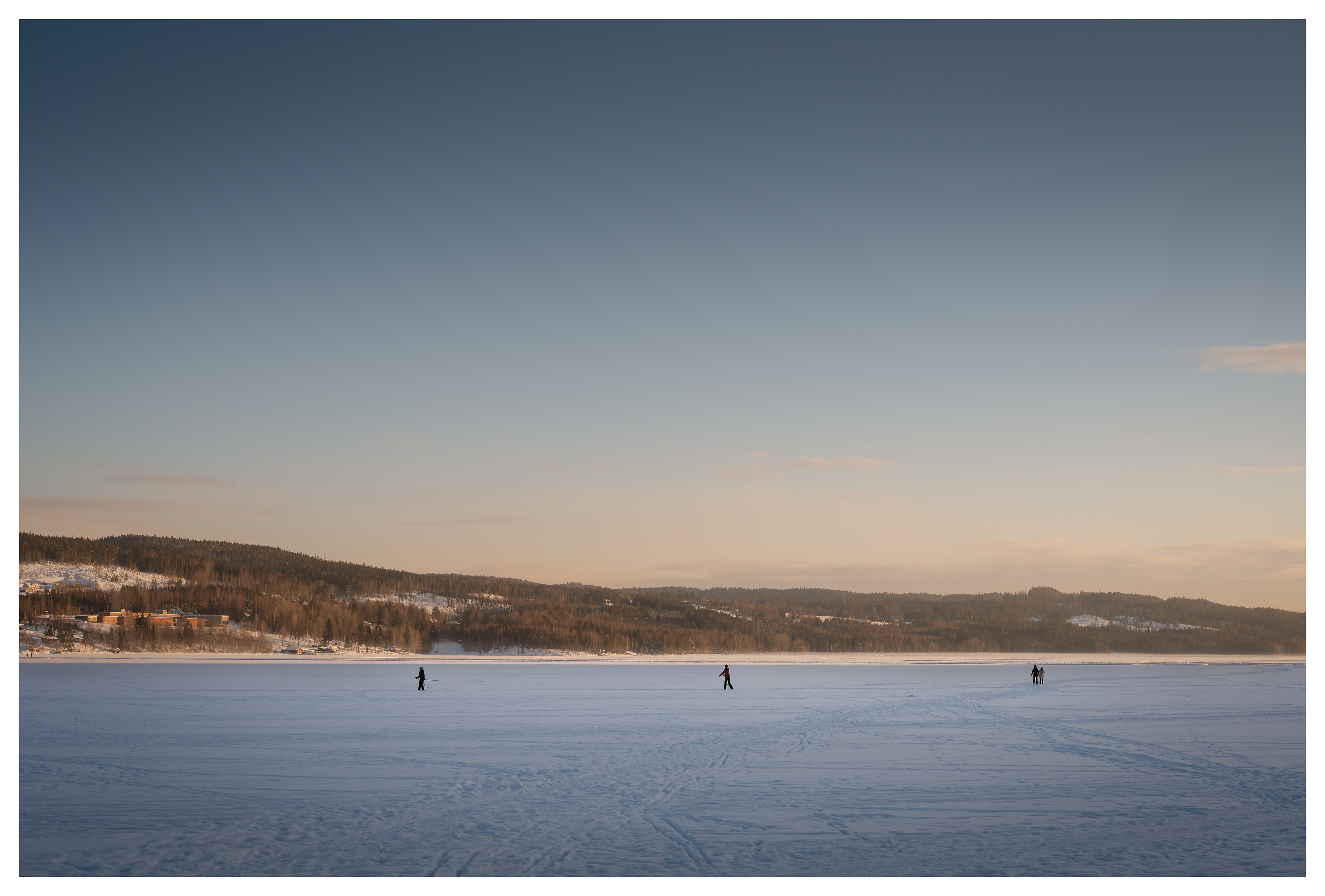 People walking separately across a frozen lake in soft winter sunset light, distant hills and tracks in the snow creating a quiet minimalist Nordic landscape.