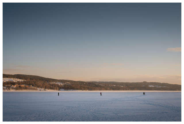 People walking separately across a frozen lake in soft winter sunset light, distant hills and tracks in the snow creating a quiet minimalist Nordic landscape.