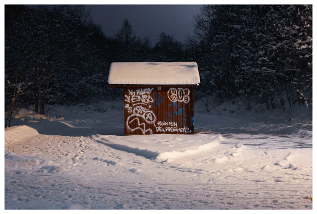Snow-covered wooden hut with graffiti in a winter forest landscape at dusk.
