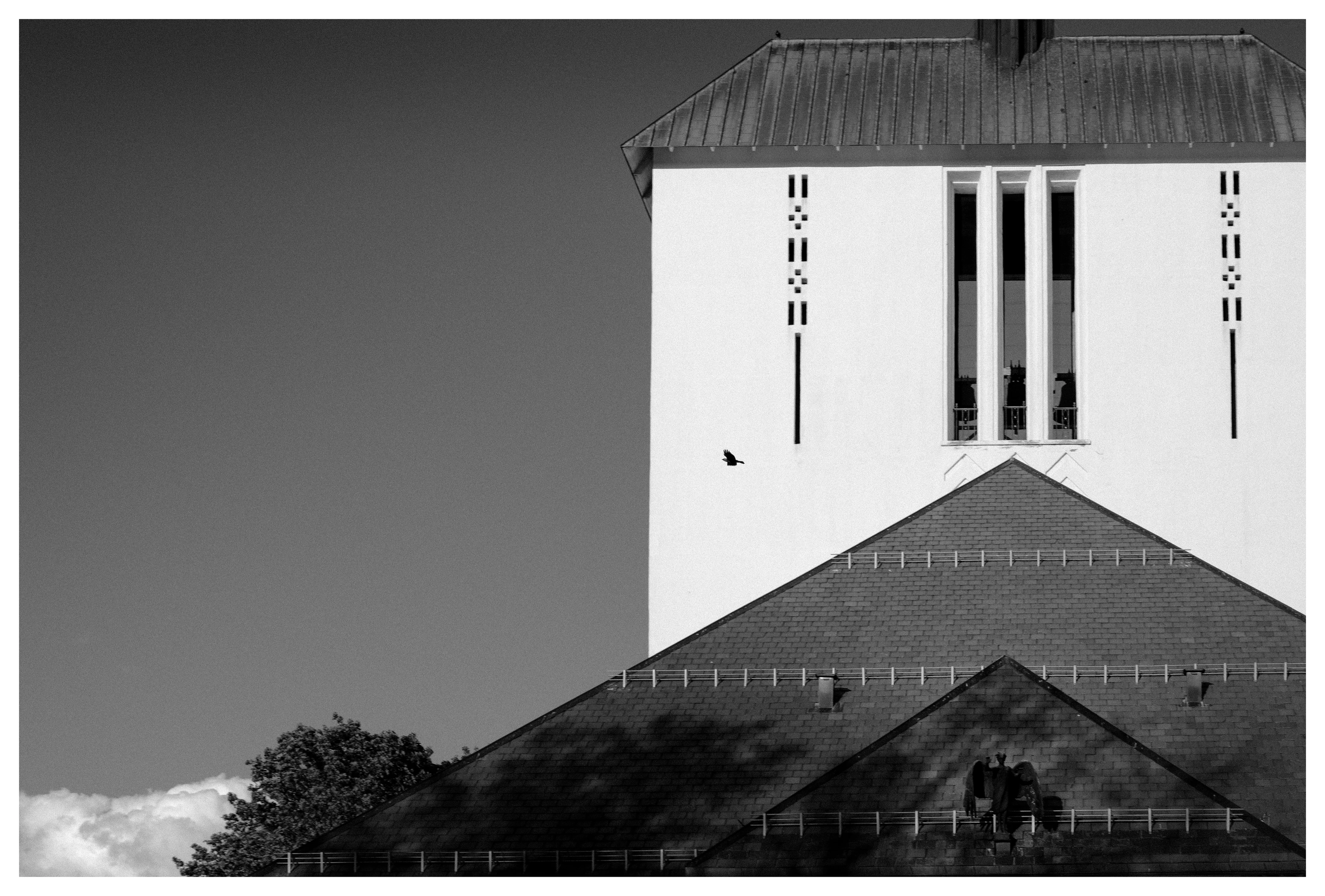 Black and white photograph of a church tower facade with geometric windows and a lone bird flying across a blank sky, emphasizing silence and scale.