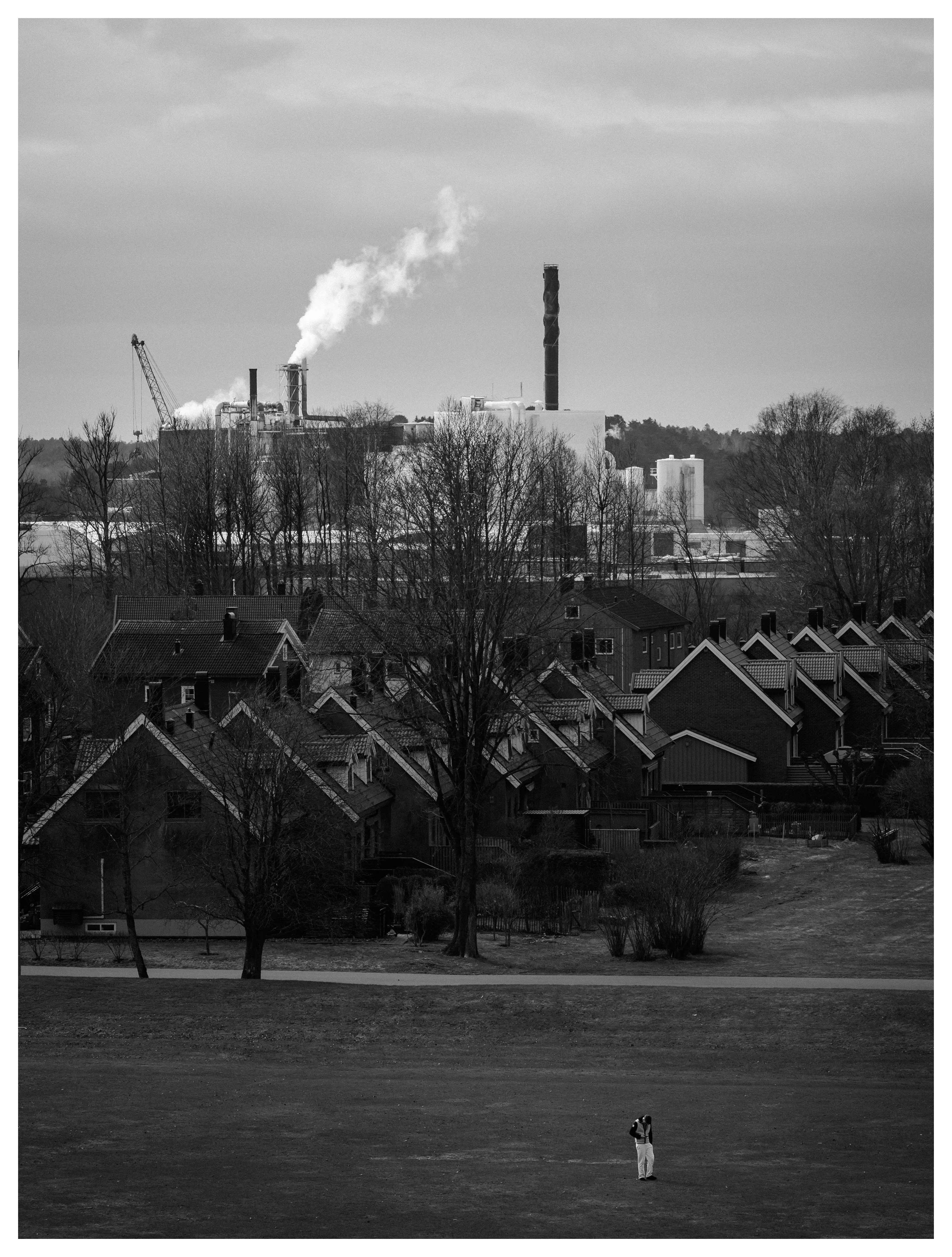 Black and white photo of a lone person standing on a field in front of suburban houses and an industrial factory with smoke rising in the background.