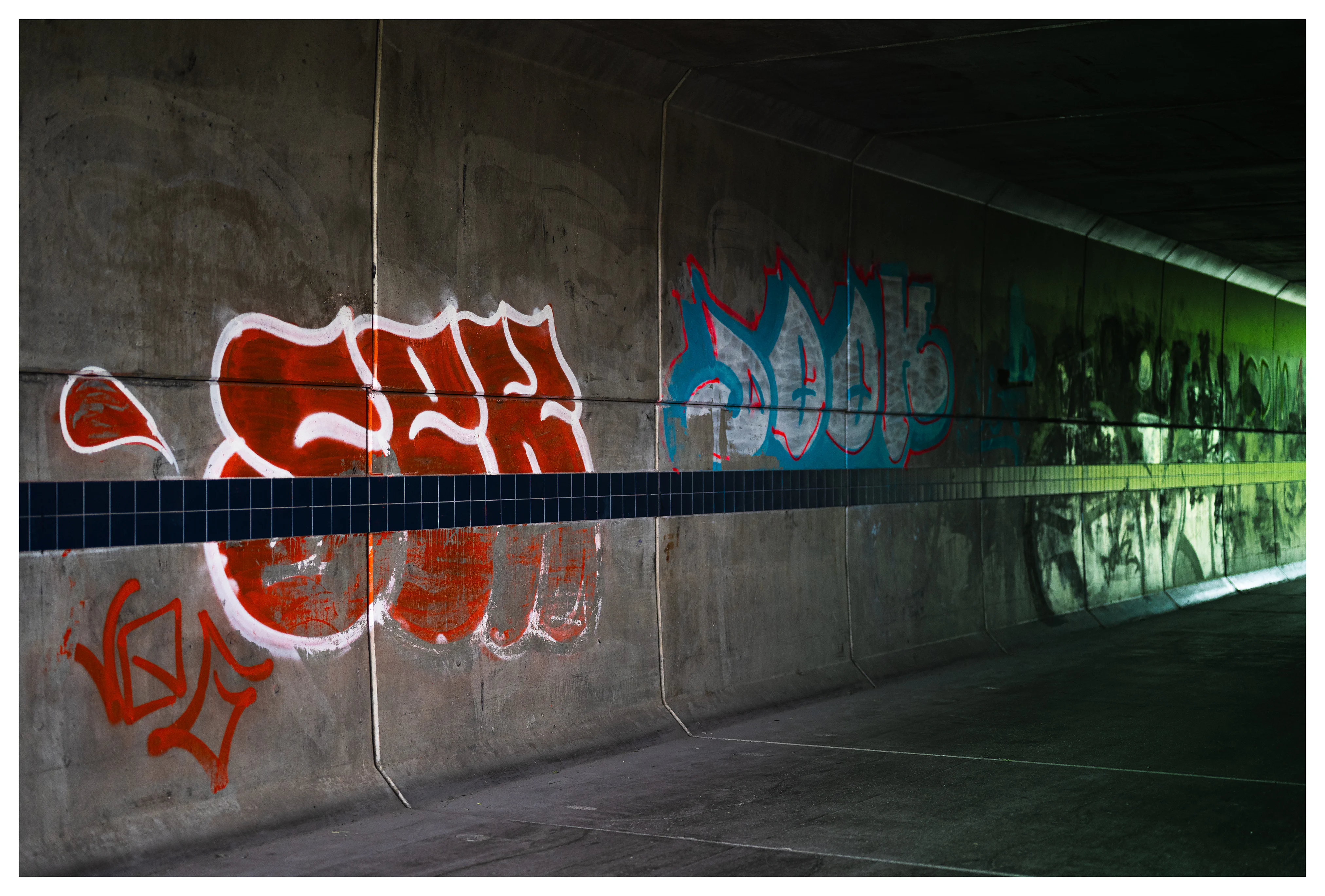Graffiti on the concrete wall of an underpass, with red and blue tags illuminated by faint green light.
