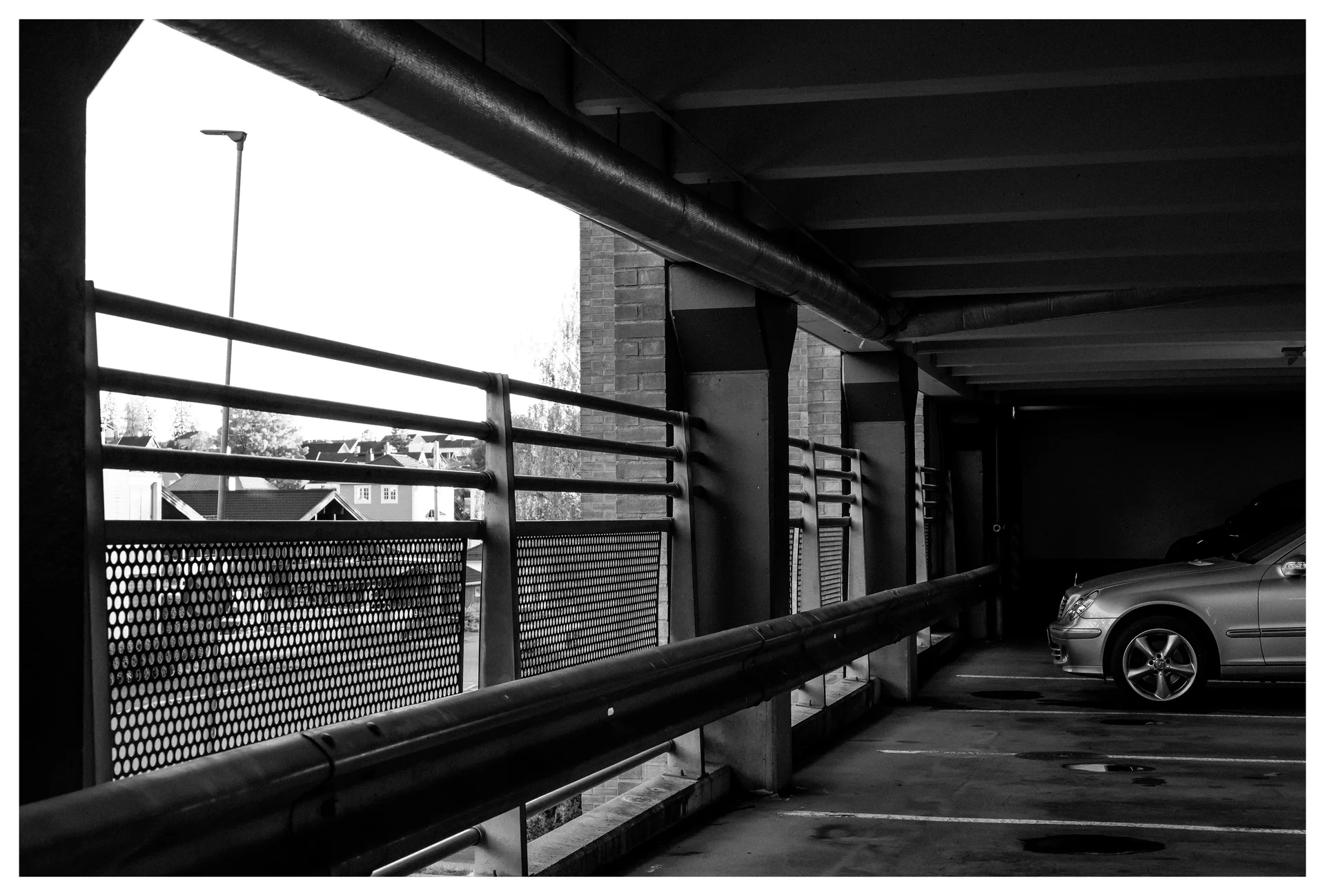 Parking garage interior on level three with railings, pipes, and a parked car in shadow.