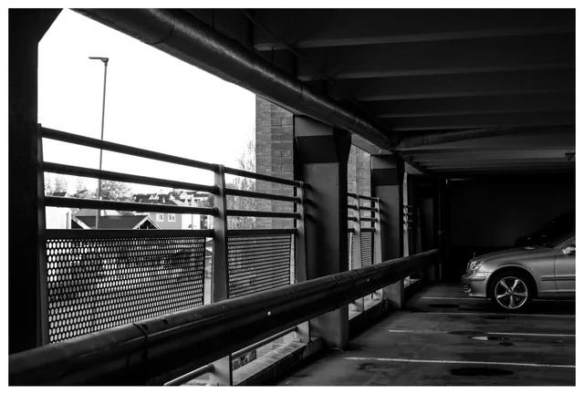 Parking garage interior on level three with railings, pipes, and a parked car in shadow.