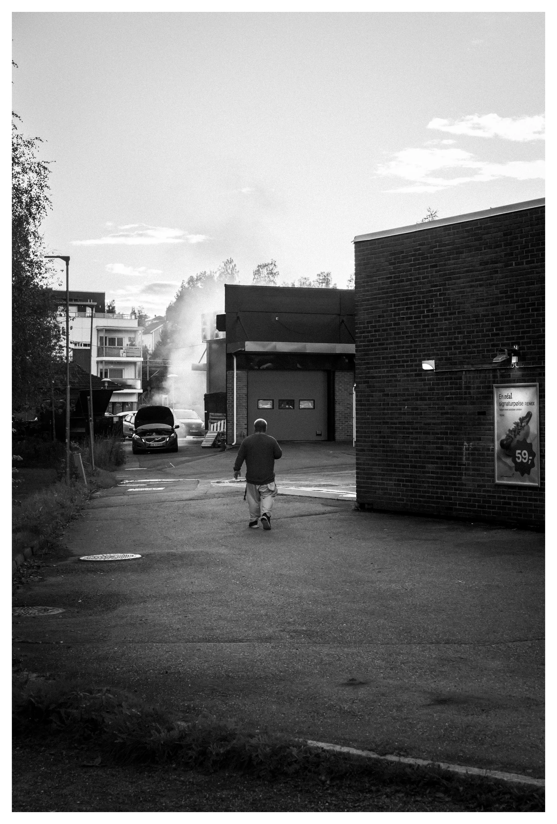 Black and white photo of a man walking down an alley toward a smoking car with its hood open near a brick building.