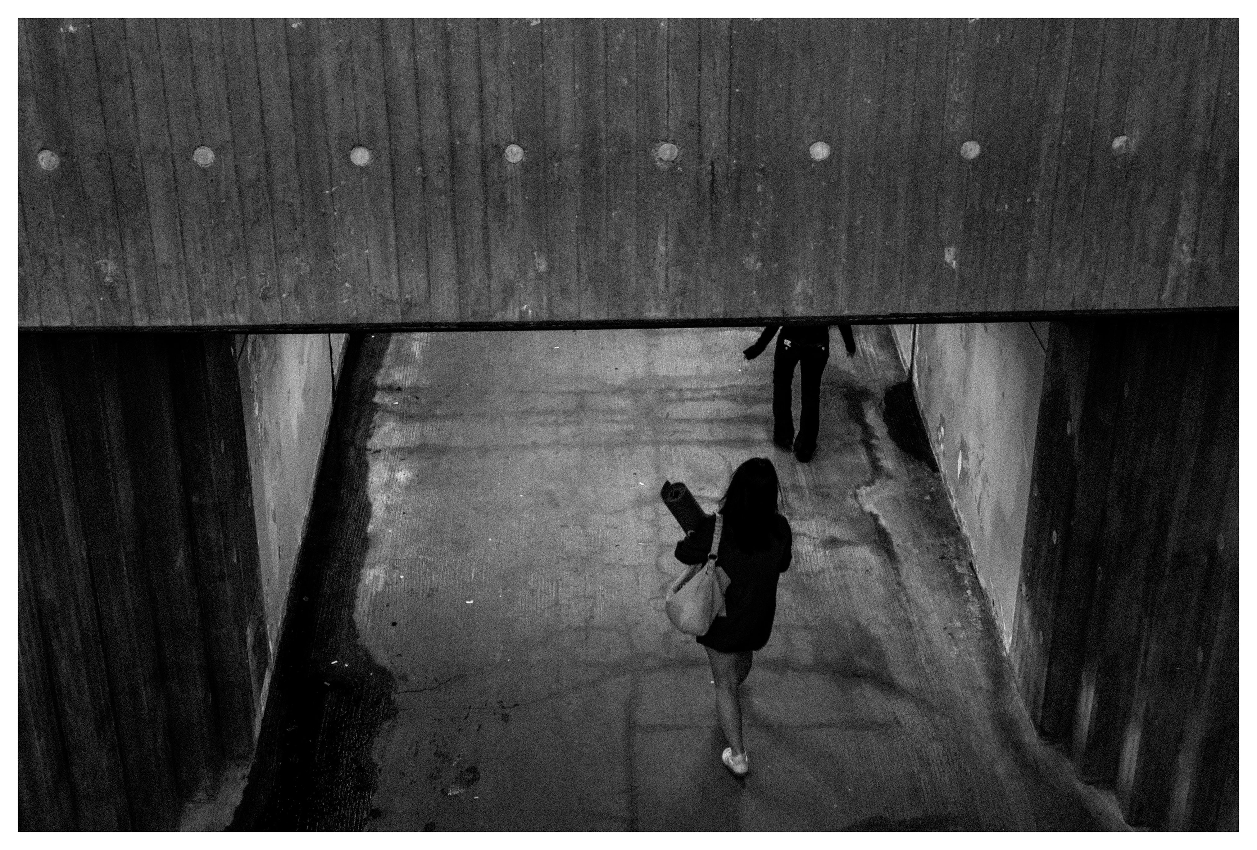 Black and white street photo of two people walking through a concrete underpass, viewed from above.