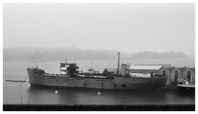 Weathered cargo ship moored in a foggy industrial harbor, black and white.