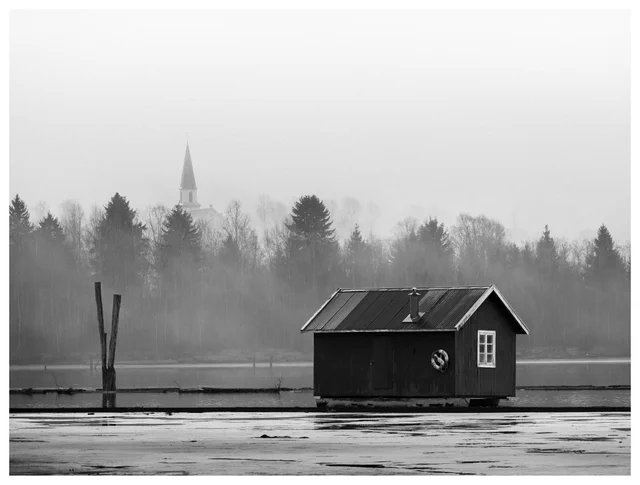 Black and white landscape with a dark wooden boathouse by a lake, fog over trees, and a church steeple in the background.