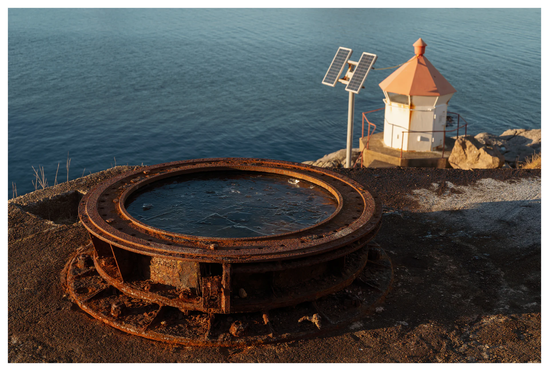 Rusted circular gun mount at Hausvikodden Fort with a small lighthouse and calm sea in the background, photographed in warm evening light.