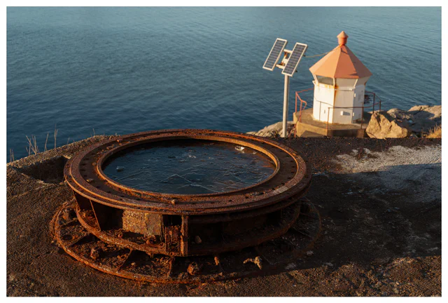 Rusted circular gun mount at Hausvikodden Fort with a small lighthouse and calm sea in the background, photographed in warm evening light.