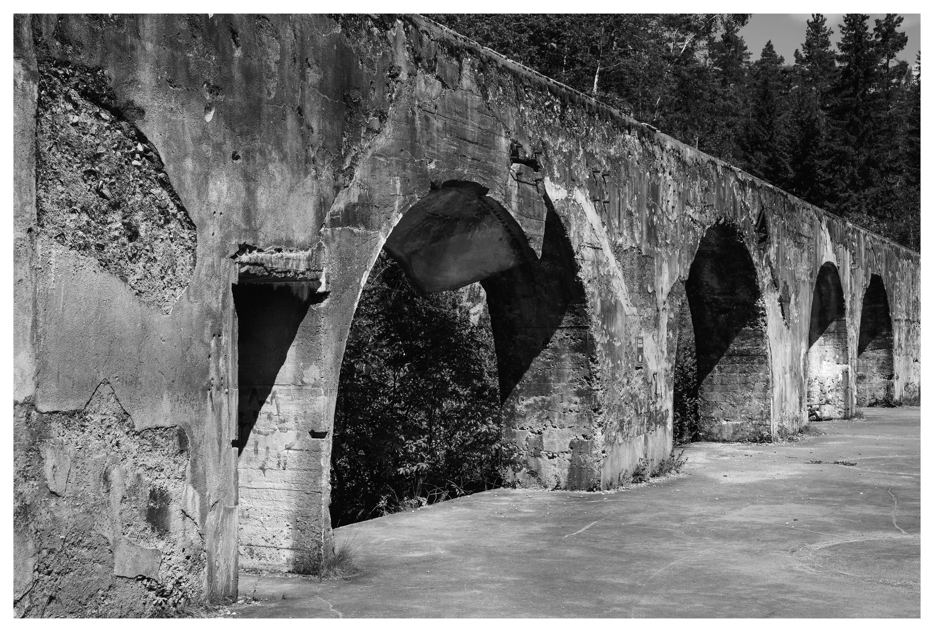 Black and white photograph of the decaying concrete arches at Kringsjå Power Station in Vennesla, showing weathered textures, deep shadows, and overgrown forest in the background.