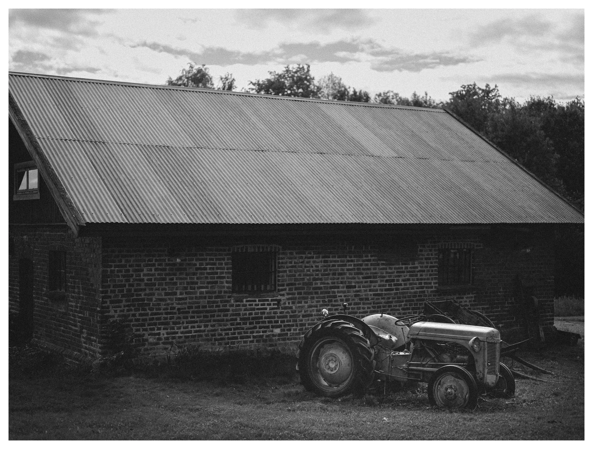 Black and white photograph of a vintage tractor parked beside an old brick farm building in the countryside.