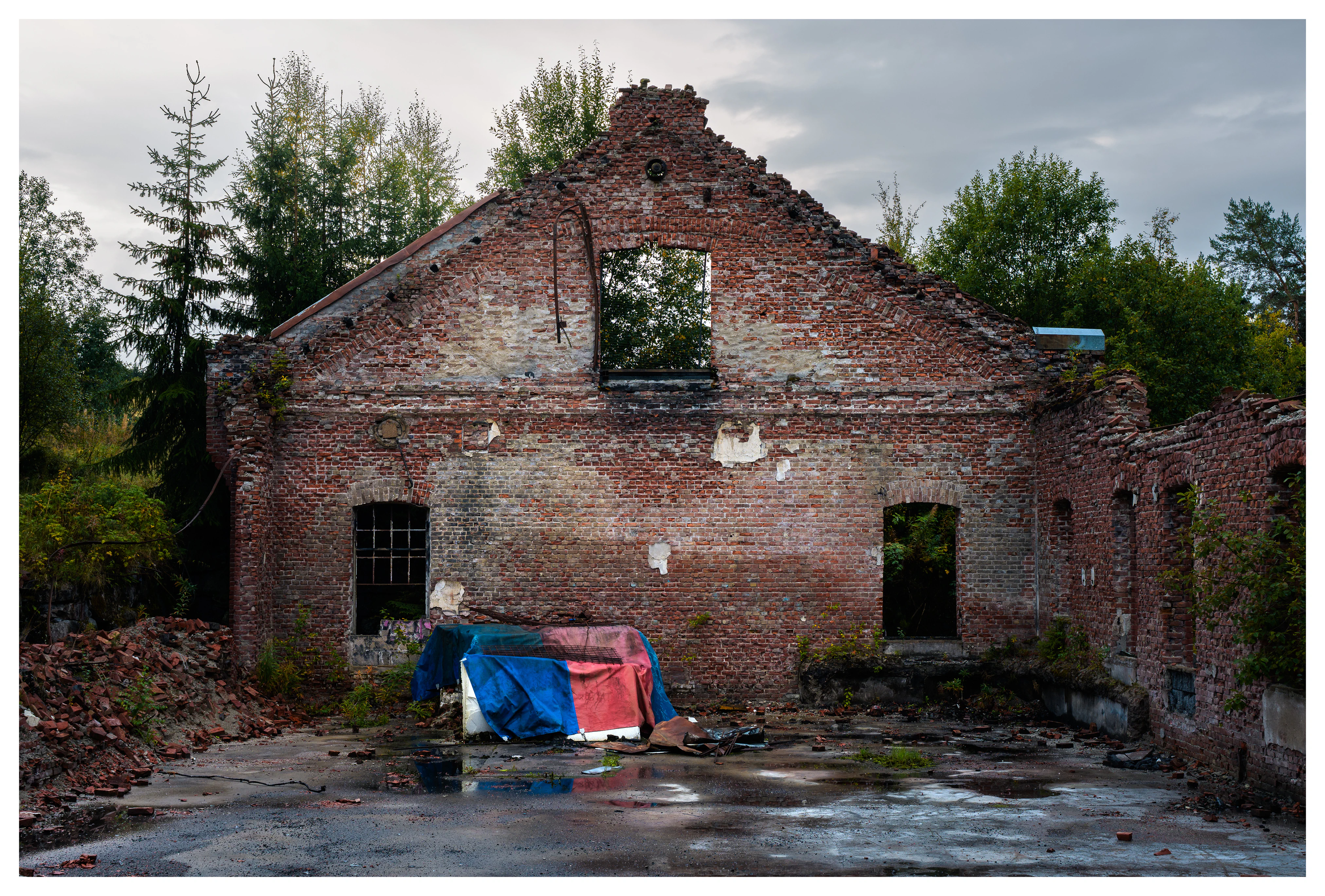 Front view of a roofless brick hall at the abandoned Mago C power plant in Eidsvoll, with broken windows, exposed walls, scattered debris, and a colorful tarp on the ground.