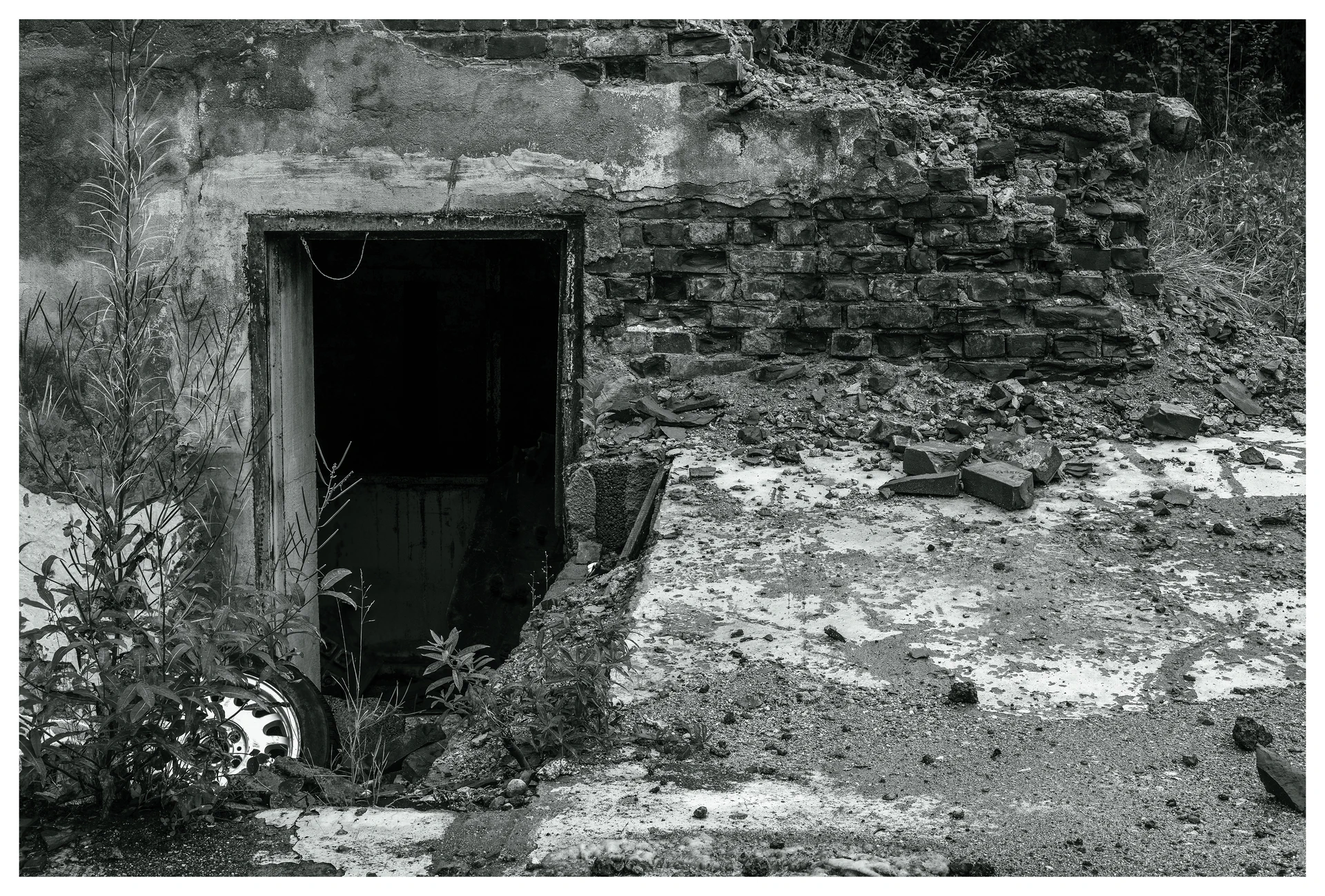 Crumbling lower entrance at the abandoned Mago C power plant in Eidsvoll, featuring a dark doorway leading underground, broken bricks, overgrown vegetation, and scattered debris.