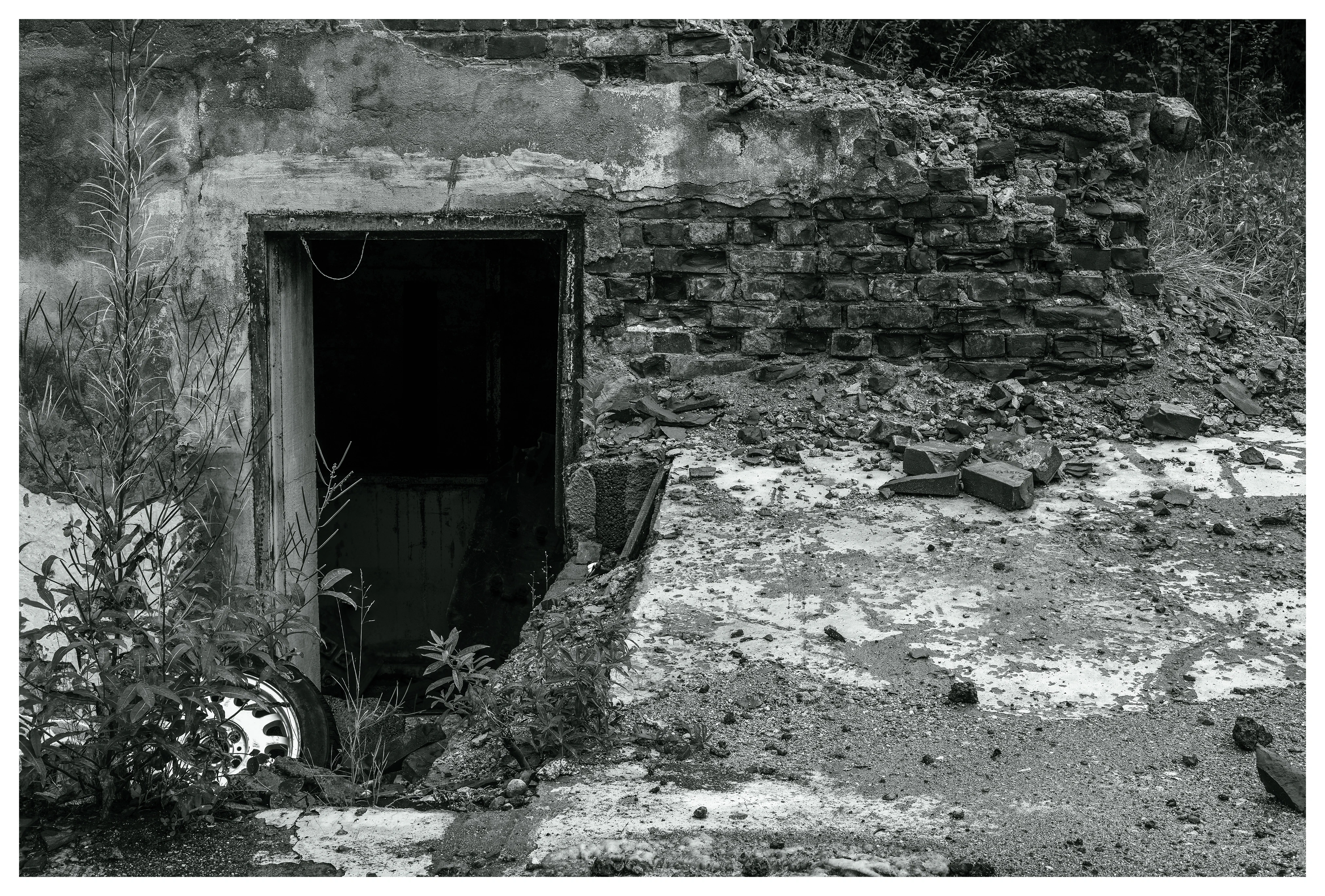 Crumbling lower entrance at the abandoned Mago C power plant in Eidsvoll, featuring a dark doorway leading underground, broken bricks, overgrown vegetation, and scattered debris.
