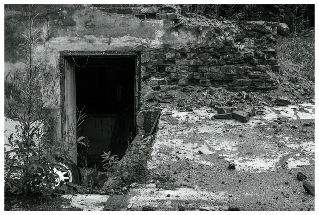 Crumbling lower entrance at the abandoned Mago C power plant in Eidsvoll, featuring a dark doorway leading underground, broken bricks, overgrown vegetation, and scattered debris.
